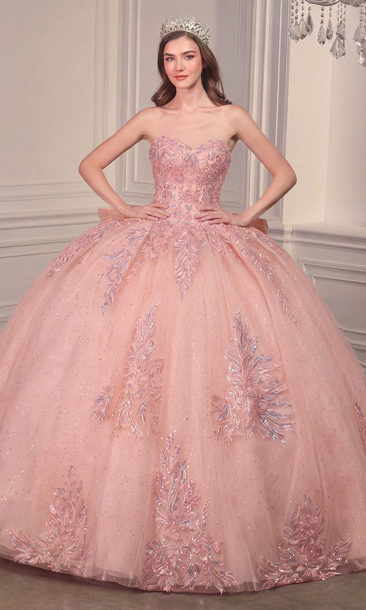 A woman in the Ladivine Strapless Quinceanera Dress 15715—sparkling pink with floral embroidery and bow detail—stands confidently, hands on hips, wearing a silver tiara in an elegant room.